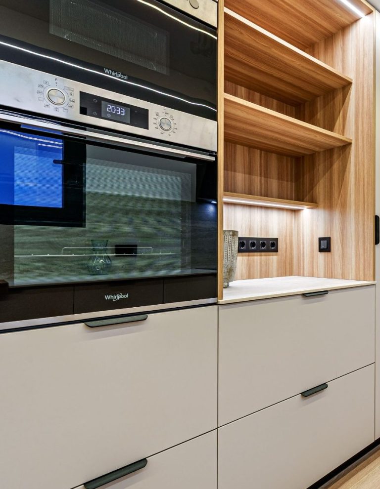 Cocina moderna con horno empotrado y estanterías de madera clara.