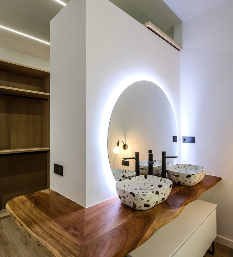 Baño moderno con lavabo de piedra y espejo iluminado en pared blanca.