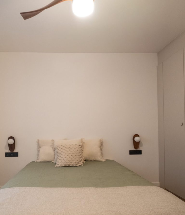 Habitación con cama doble, paredes blancas y lámpara de techo. Cómodas mesitas a los lados.