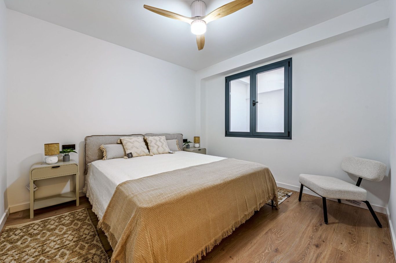 Dormitorio luminoso con cama doble, mesita de noche y sillón. Suelo de madera clara.