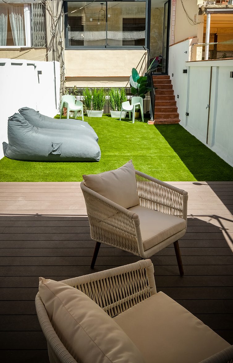 Jardín con césped, sillones y un pouf, rodeado de plantas y escaleras al fondo.
