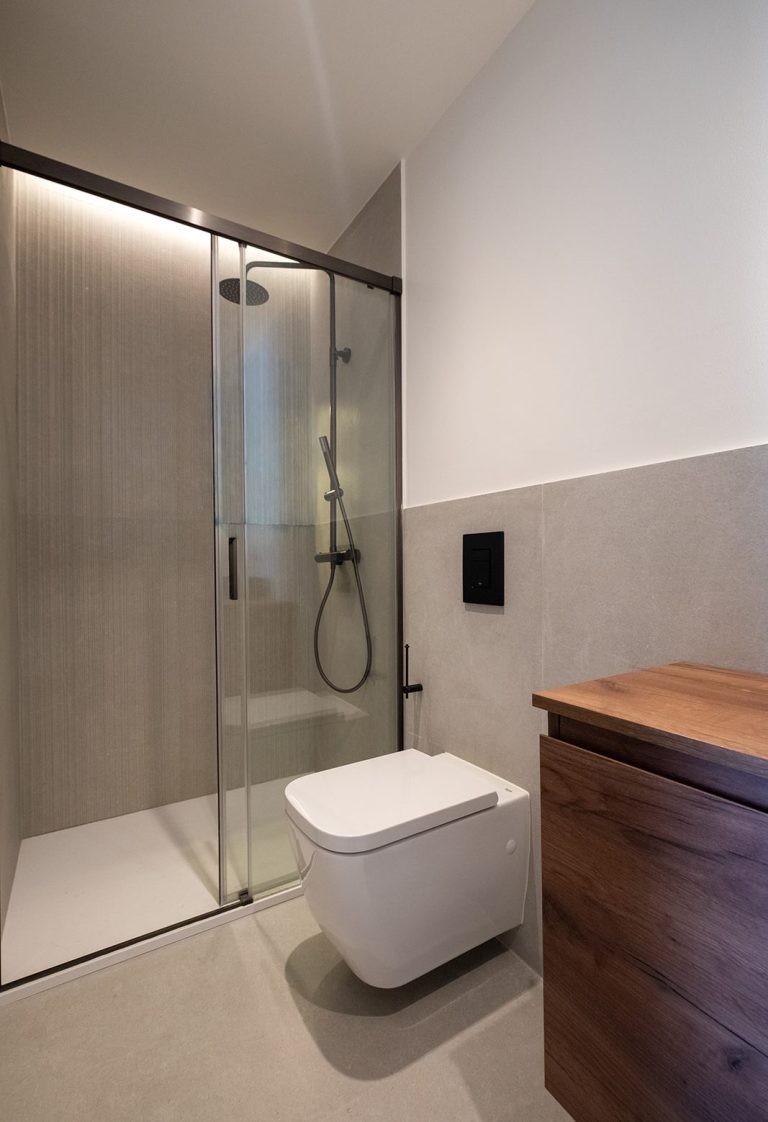 Baño moderno con ducha de vidrio, inodoro blanco y mueble de madera.