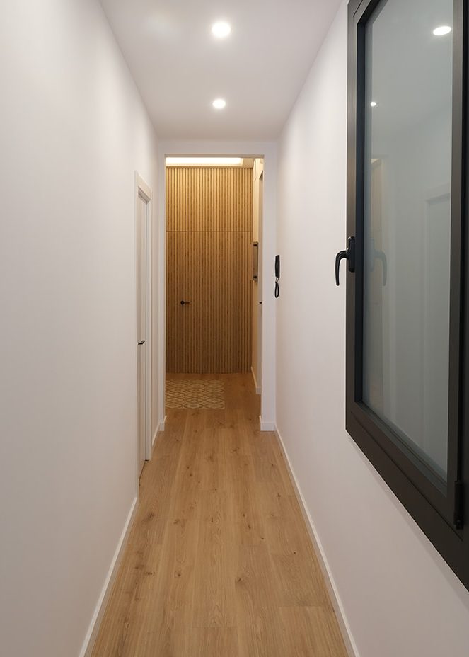 Pasillo moderno y luminoso con paredes blancas y suelo de madera clara. Puerta de madera al fondo.