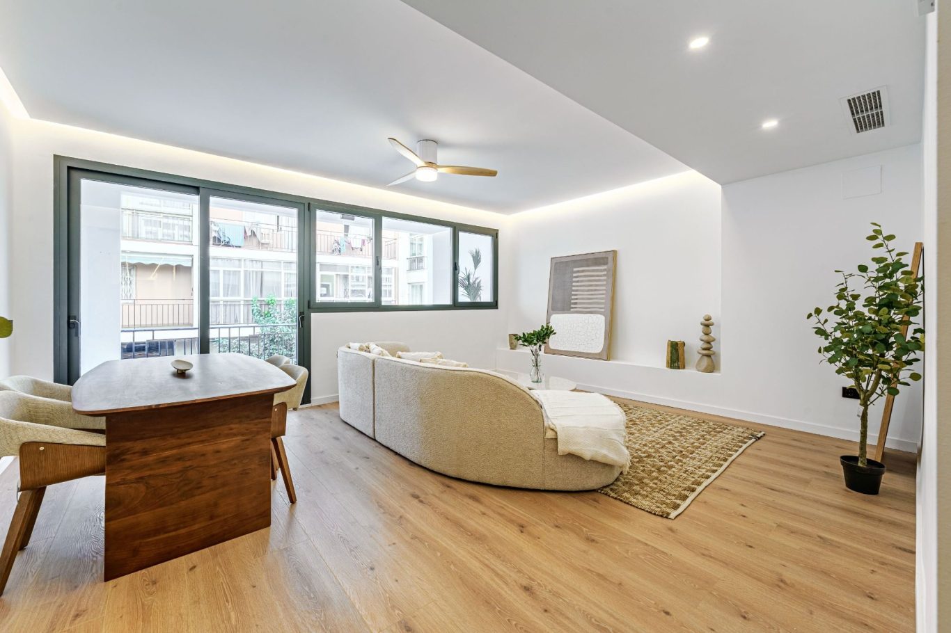 Sala de estar moderna con sofá, mesa y plantas, iluminada y con ventanas grandes.