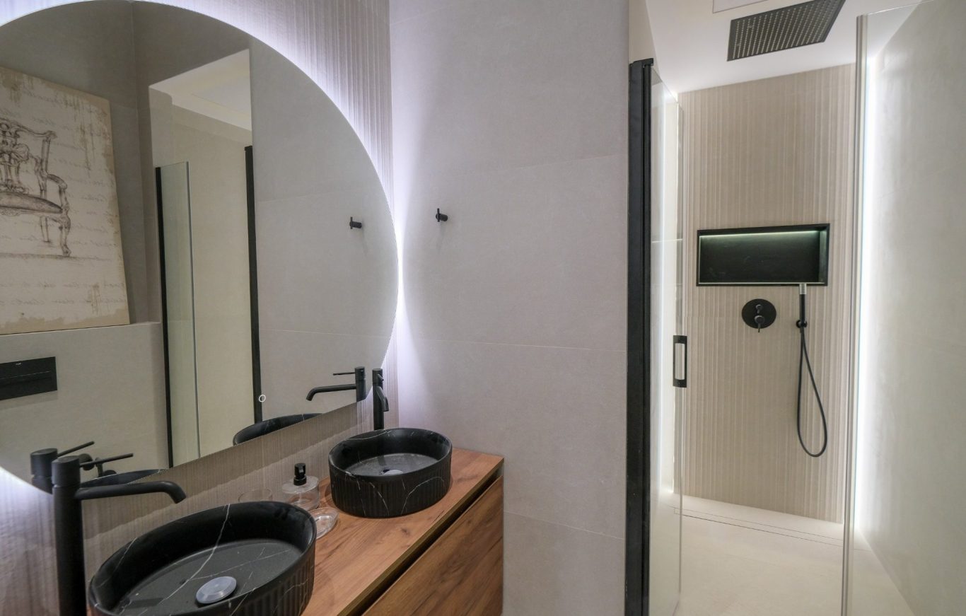 Baño moderno con lavabo de piedra, espejo circular y ducha con iluminación suave.